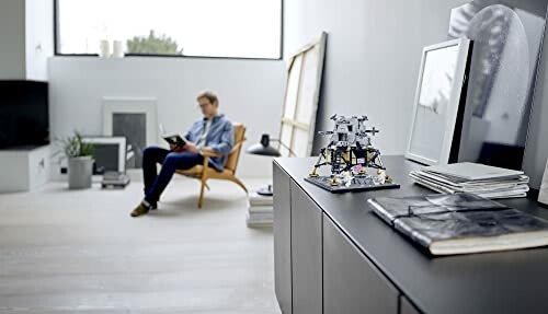 Modern living room with a person reading and a LEGO model on a cabinet.