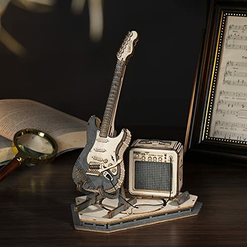 Wooden model of a guitar and amplifier on a desk with a magnifying glass and sheet music in the background.